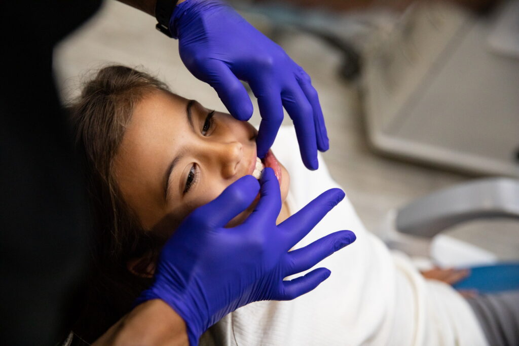 A child is seated in a dental chair, smiling as a dentist, wearing purple gloves, examines her teeth. The setting is a dental office, conveying a friendly and professional atmosphere.