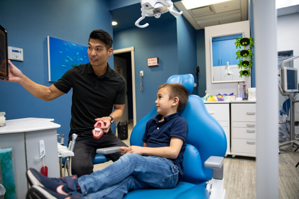 A friendly dental professional shows a child a dental model while seated in a bright, modern office with dental equipment in the background. The child looks engaged and attentive. A cheerful 123Dentist Smiley is present, adding a friendly atmosphere to the scene.