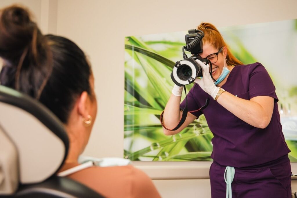 A dental professional in purple scrubs is taking a photograph of a patient seated in a dental chair. The background features a calming green plant design, emphasizing a friendly and comfortable environment.