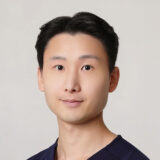 A professional portrait of a man with dark hair, wearing a navy scrub top. He has a neutral expression and a light background, suitable for a healthcare setting.