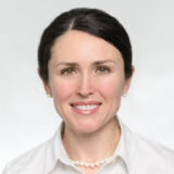 A smiling woman with dark hair and a white blouse appears against a light background. She looks approachable and friendly, exuding a sense of professionalism.