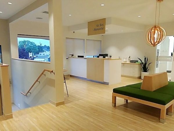 Modern dental office waiting area with light wood flooring, green cushioned seating, a front desk, and natural lighting. The layout suggests a comfortable space for patients seeking a dentist near you.