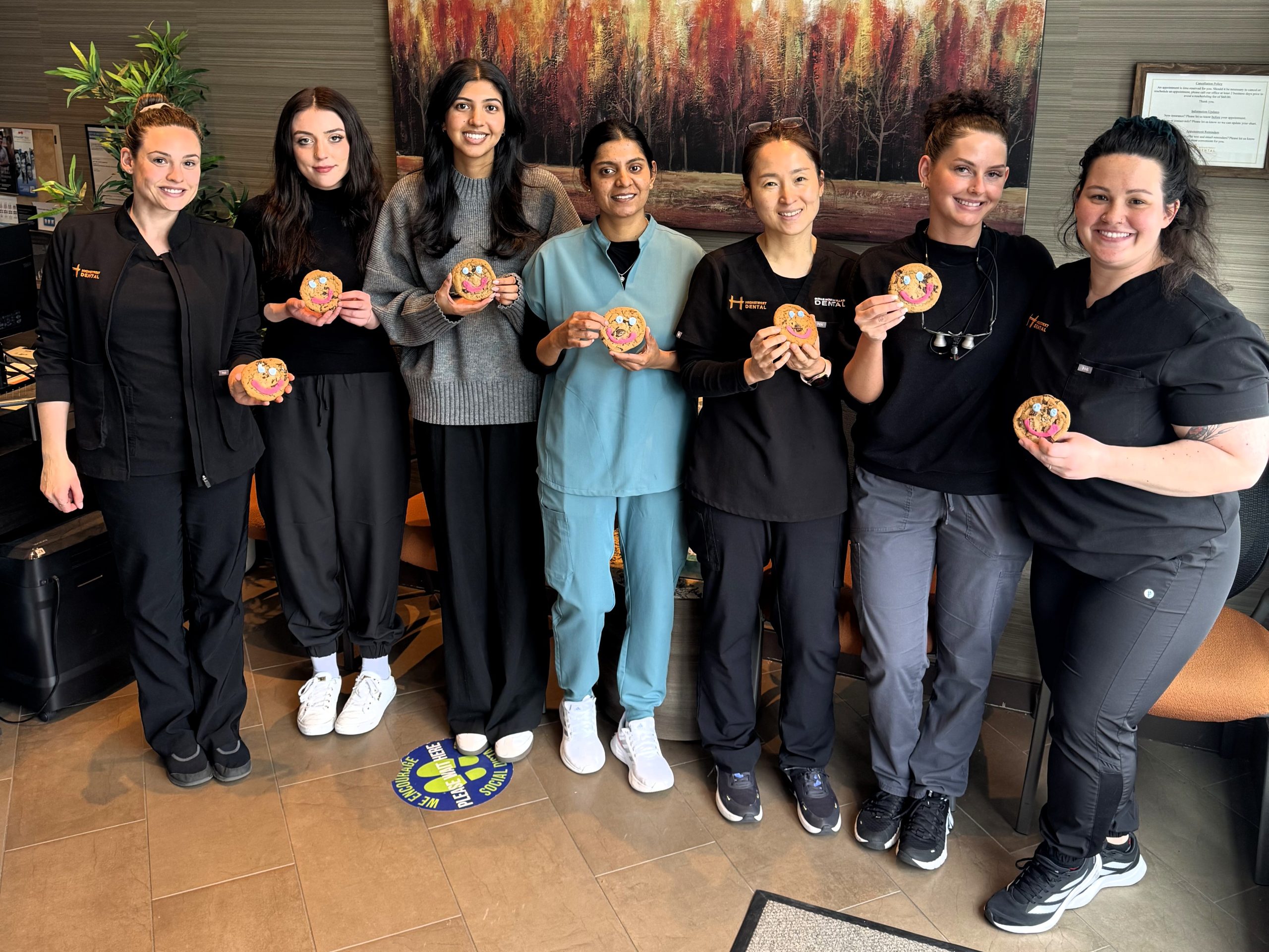 A group of seven dental professionals stands together, smiling and holding cookies in their hands. They are dressed in various uniforms, showcasing a friendly and welcoming atmosphere in a dental office. In the background, there is a colorful abstract artwork. The 123Dentist Smiley is also present, adding a cheerful touch.