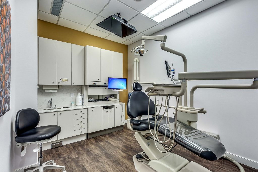 A modern dental office featuring a dental chair with overhead lighting, a computer screen, and storage cabinets against a wall. The room has a neutral color scheme with wooden flooring and a minimalist design.