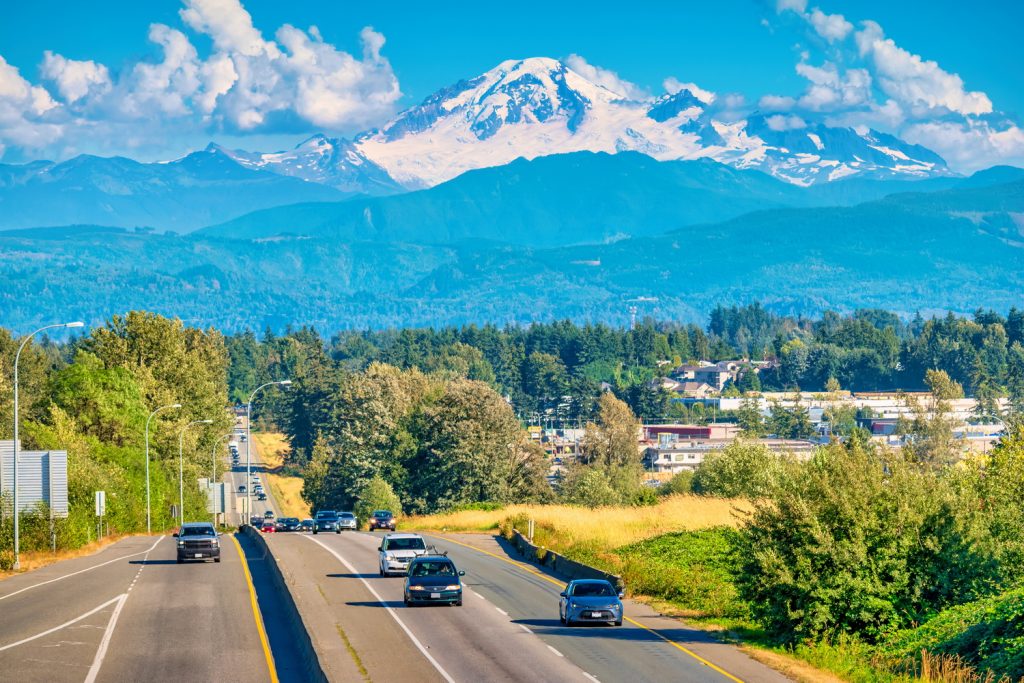 A scenic view of a highway bordered by lush trees and fields, leading toward snow-capped mountains in the background. A cheerful blue smiley face is superimposed on the image, adding a whimsical touch to the natural landscape.