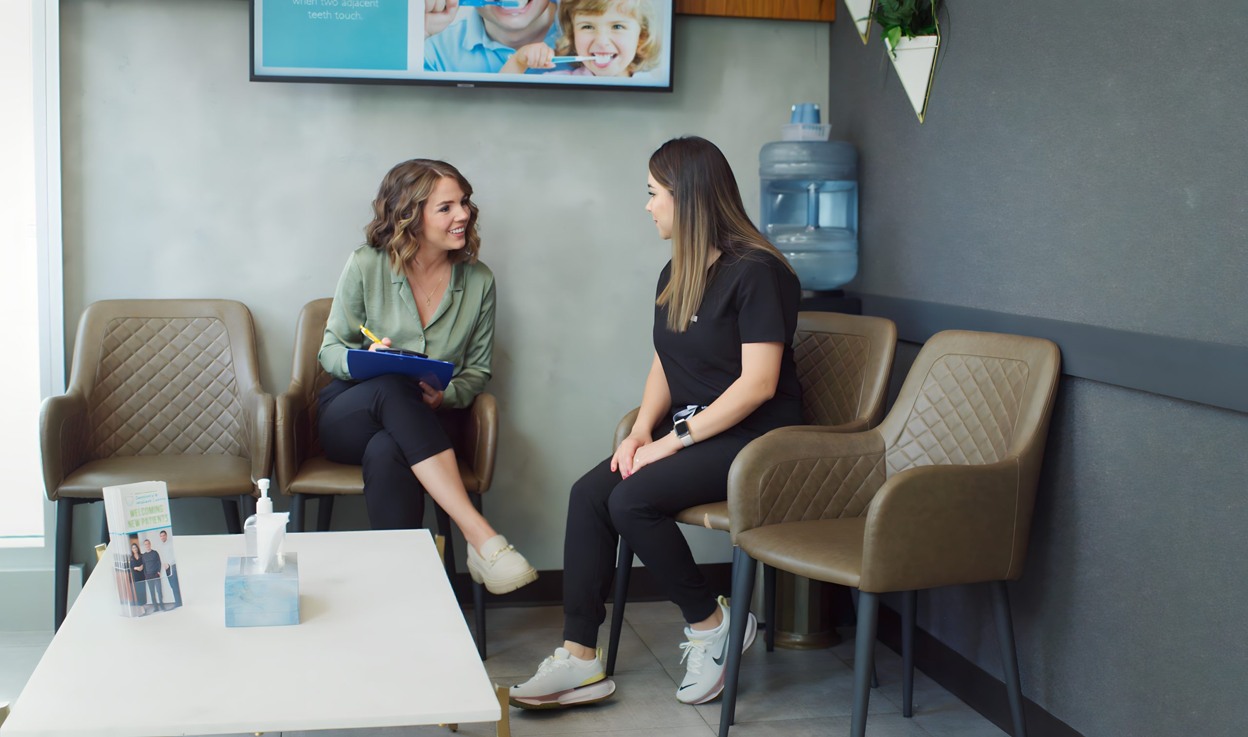Two women are sitting in a waiting area, engaged in conversation. One woman holds a clipboard, while the other listens attentively. The space features modern furniture and a water cooler in the background.