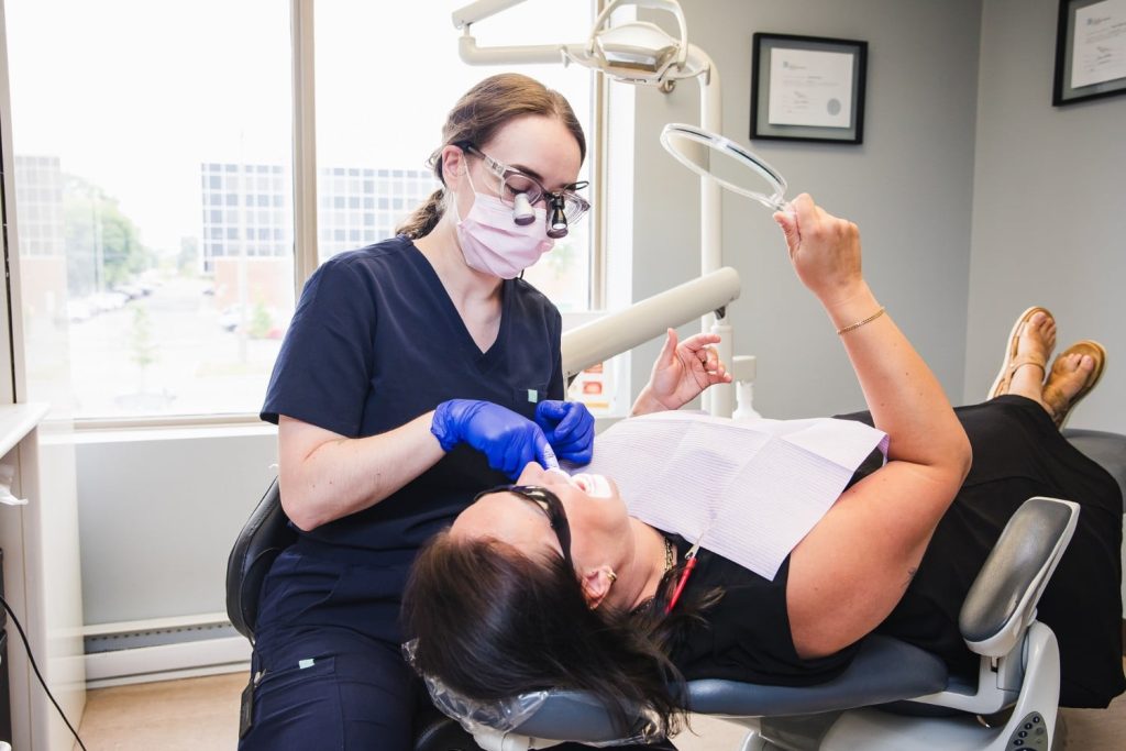 A dental hygienist works on a patient in a modern clinic, using tools to provide care. The patient lies back in the dental chair, wearing protective eyewear, while a large window reveals a view outside. The friendly 123Dentist Smiley adds a cheerful atmosphere to the setting.