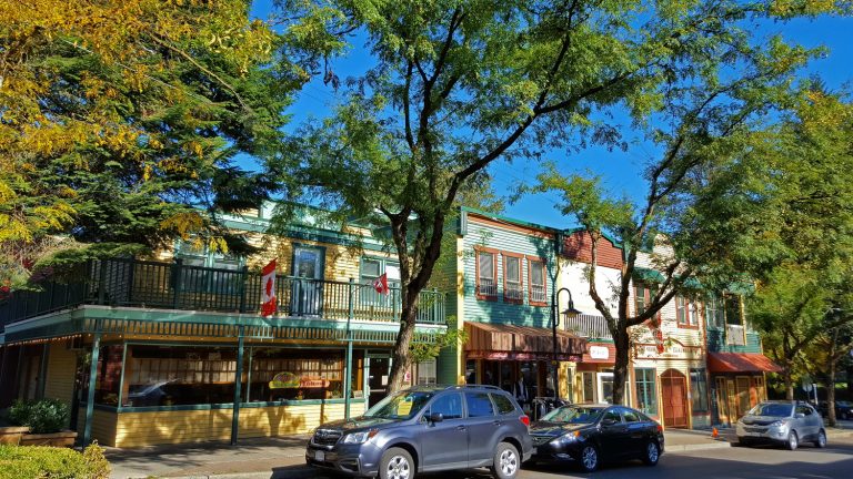 A vibrant street scene featuring colorful buildings with balconies, trees providing shade, and parked cars along the curb, all under a clear blue sky with a smiling face graphic overlay.
