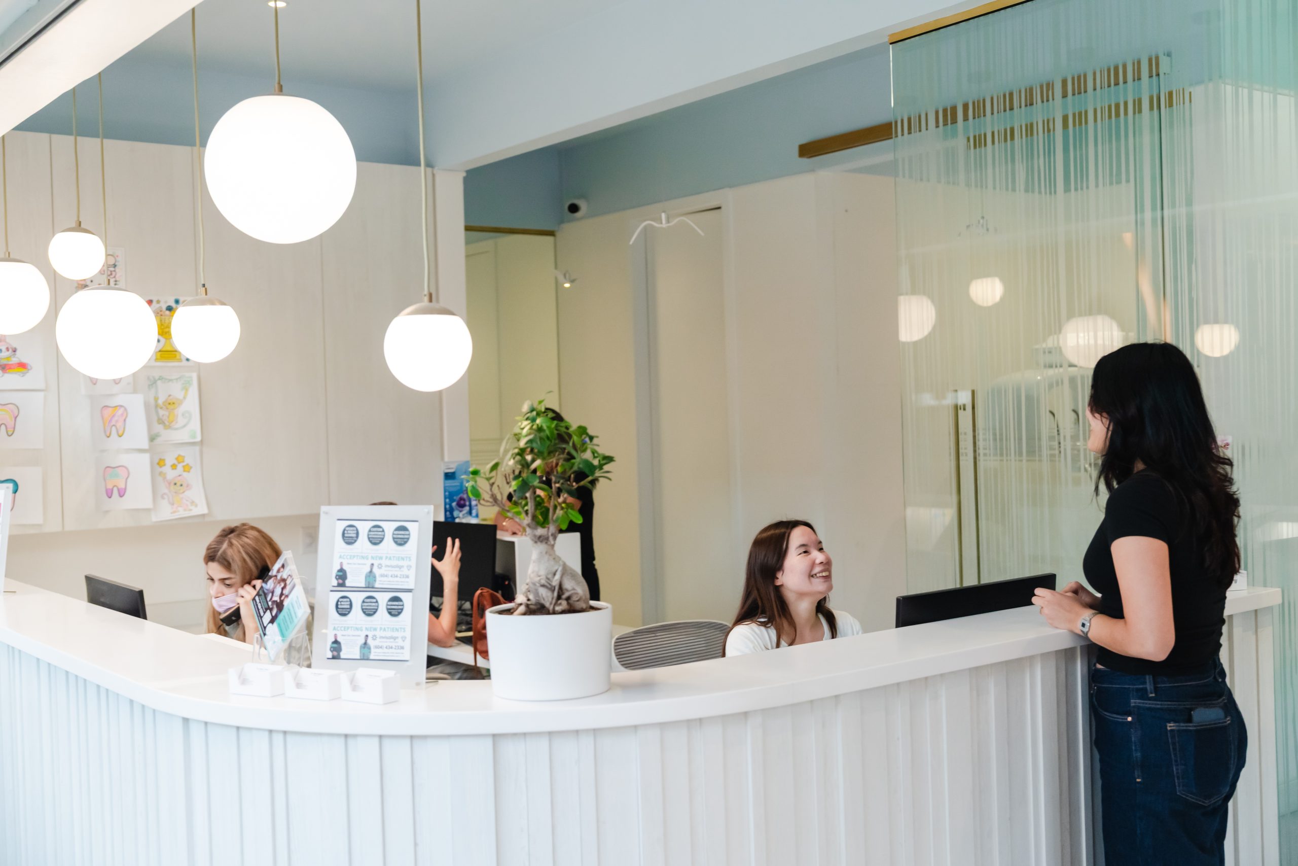 A friendly dental office reception area with two staff members engaged at the front desk while a visitor speaks with another staff member. The space is bright and welcoming, featuring modern lighting and greenery for a pleasant atmosphere. The 123Dentist Smiley is also present to add cheer.