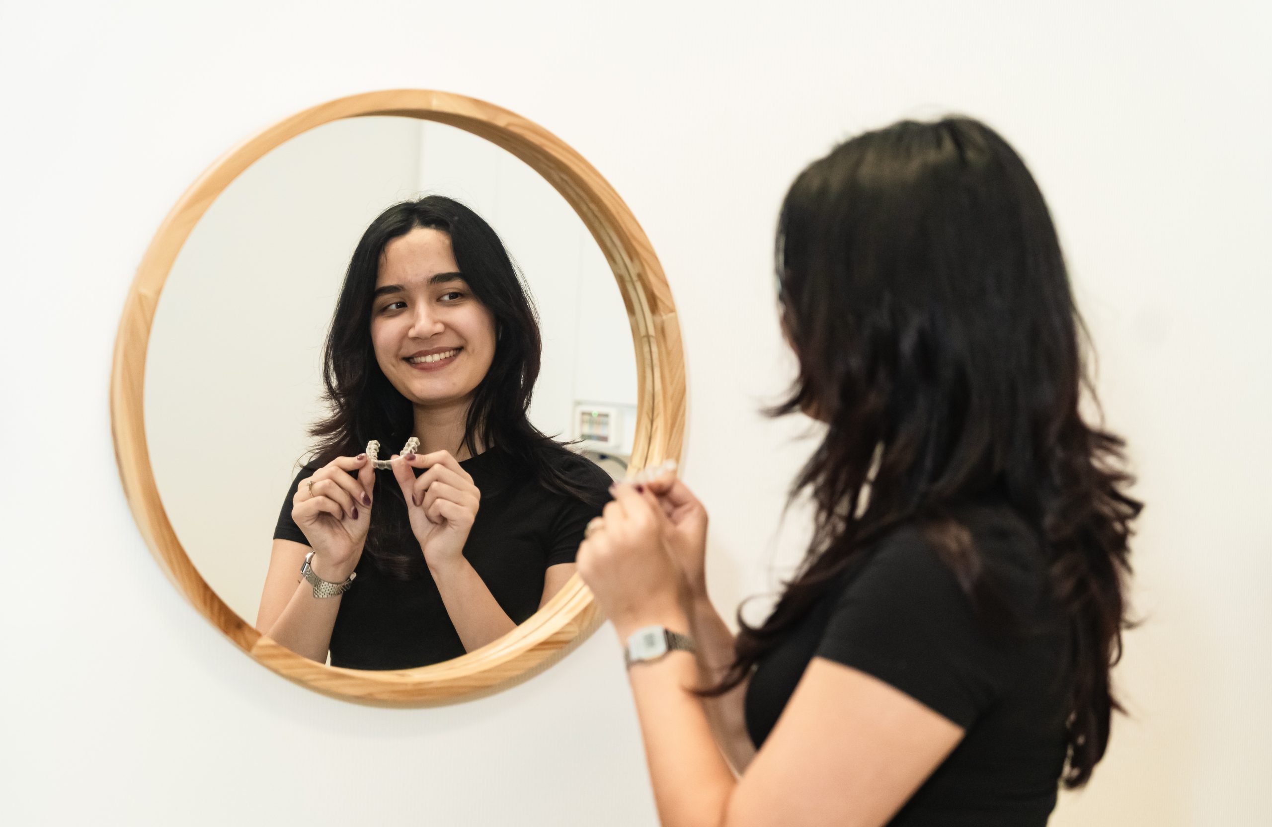 A woman with long hair smiles at her reflection in a round, wooden-framed mirror, holding small objects in her hands. She is wearing a black shirt and appears to be in a bright, minimalistic setting.