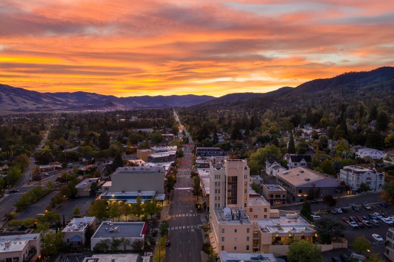 A scenic aerial view of a suburban town at sunset, with vibrant colors in the sky. A large, blue smiley face graphic is superimposed over the landscape, adding a whimsical touch to the serene setting.