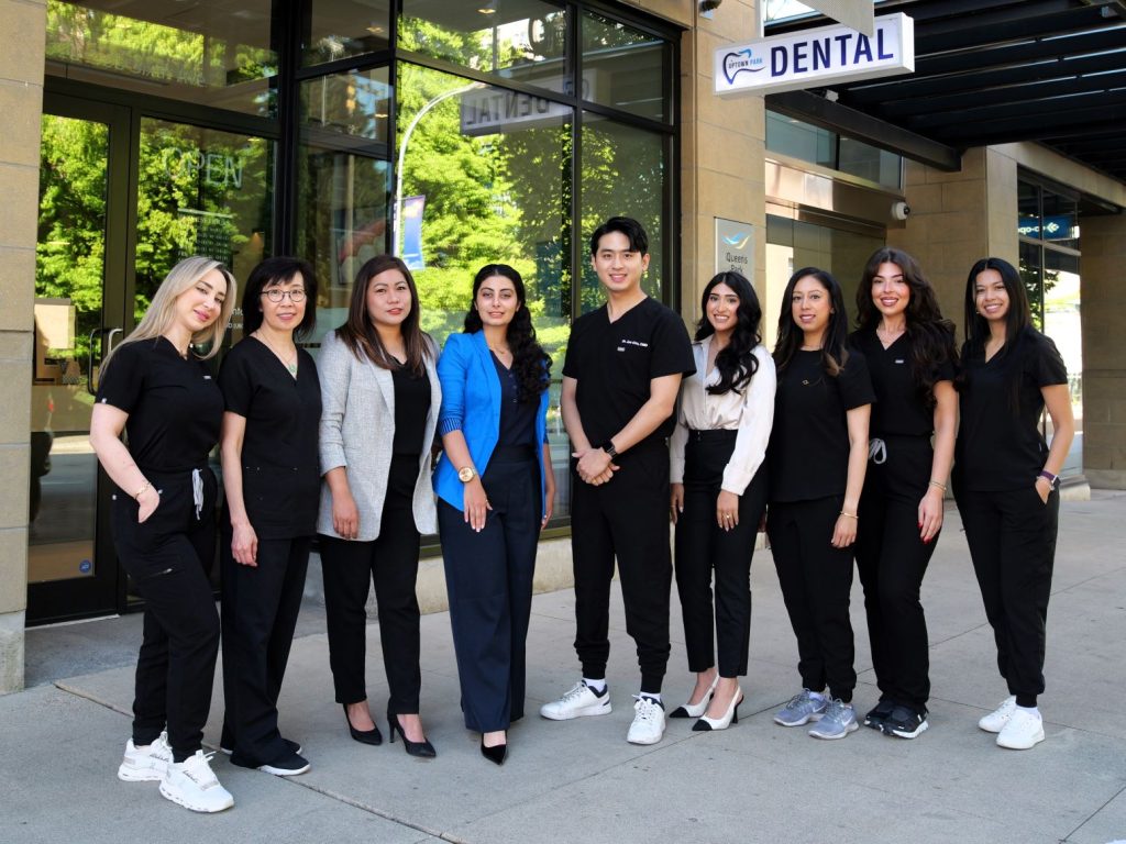 A diverse group of dental professionals stands outside a dental clinic, dressed in black scrubs. They smile and pose together, showcasing team spirit and approachability. In the background, the clinic's sign is visible.