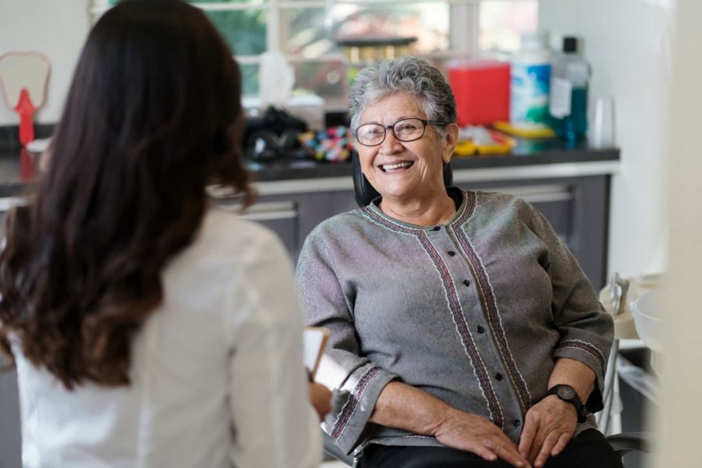 A smiling older woman sits in a dental office, engaging with a dental professional. The setting is bright and welcoming, reflecting a friendly atmosphere. The 123Dentist Smiley adds a cheerful touch to the scene, promoting a positive dental experience.