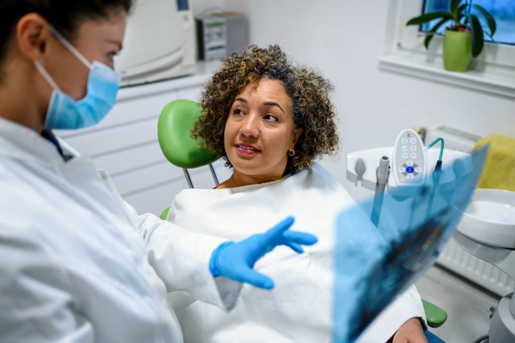 A dentist in a white coat discusses an X-ray with a smiling patient in a dental chair. The patient, wrapped in a dental bib, looks attentive as they engage in conversation.