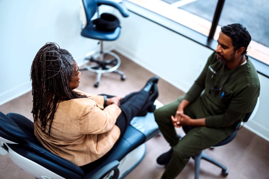 A dental professional converses with a patient seated in a dental chair in a modern office. The setting is well-lit, featuring dental equipment and a calm atmosphere, indicating a friendly and informative consultation.