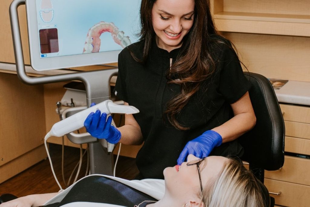A dental professional smiles at a patient reclining in a dental chair, holding a dental tool. A monitor in the background displays dental imagery, creating a welcoming and informative environment. The scene conveys a supportive dental experience.