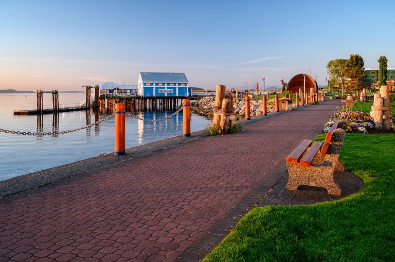 A scenic waterfront path lined with benches, leading to colorful buildings and docks under a clear blue sky. The calm water reflects the surroundings, creating a tranquil atmosphere.