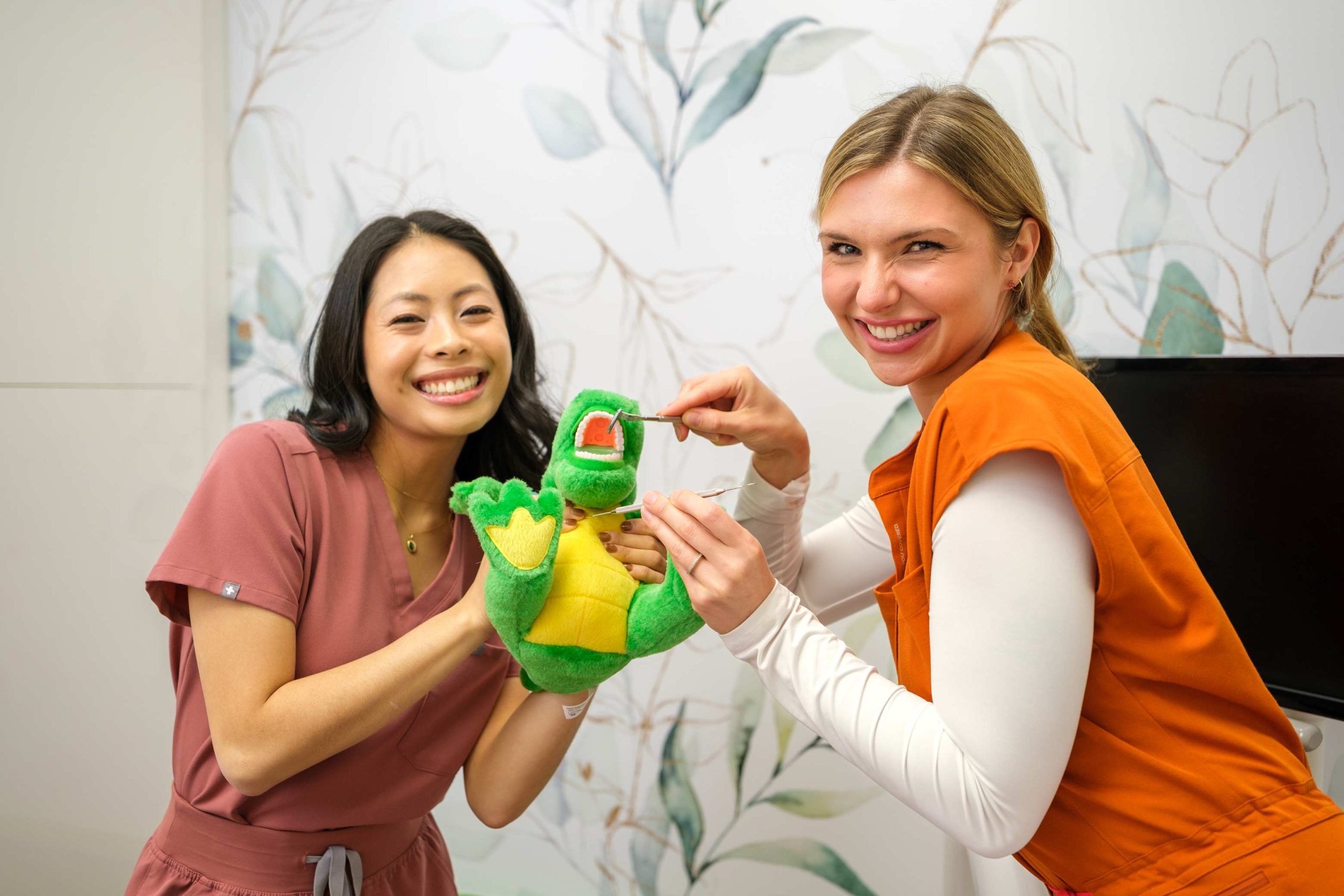 Two women, one in a pink scrubs and the other in an orange top, smile while holding a green dinosaur puppet. They appear to be in a bright, cheerful room with floral decor, promoting a friendly dental environment. The 123Dentist Smiley adds a playful touch to the scene.