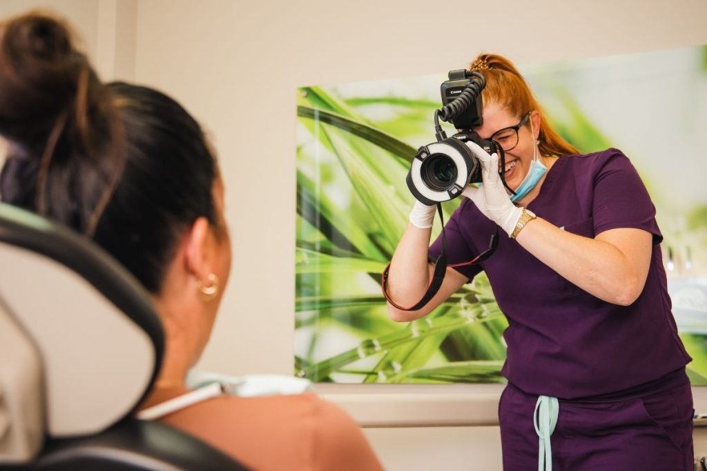 A dental professional in purple scrubs is taking a photograph of a patient seated in a dental chair. The background features a calming green plant design, emphasizing a friendly and comfortable environment.