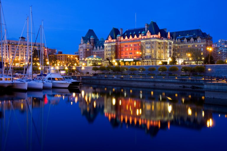 Waterfront view of a lit-up, grand building at dusk, reflecting in the calm water, with docked boats on the left and a smiling blue emoji in the sky.