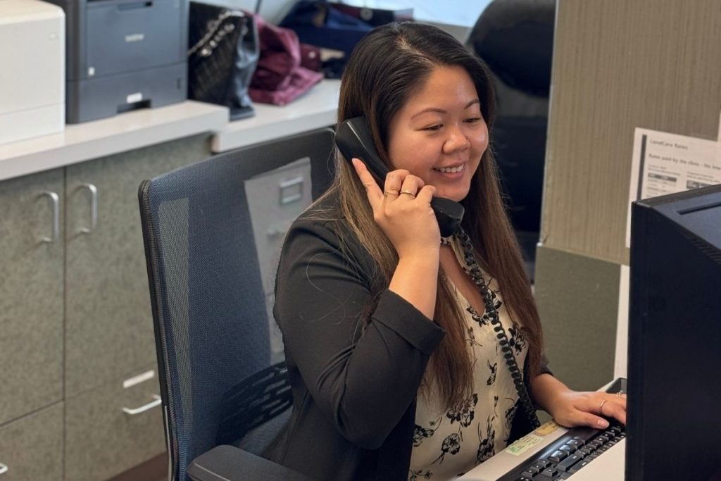 A receptionist wearing a black blazer smiles while speaking on the phone at her desk. She is engaged with a computer, showcasing a friendly and professional atmosphere in a dental office. The 123Dentist Smiley is not included in this image.