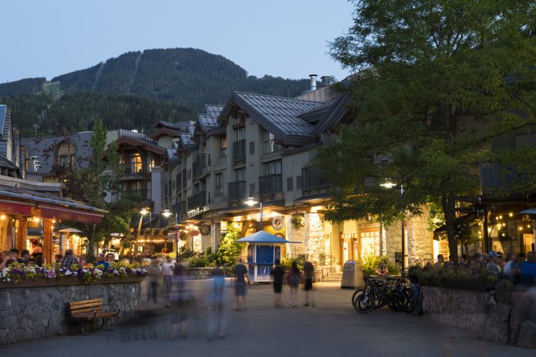 A bustling evening scene in a village with people walking, illuminated shops, and a backdrop of mountains. A blue circle with a smiling face hovers above.